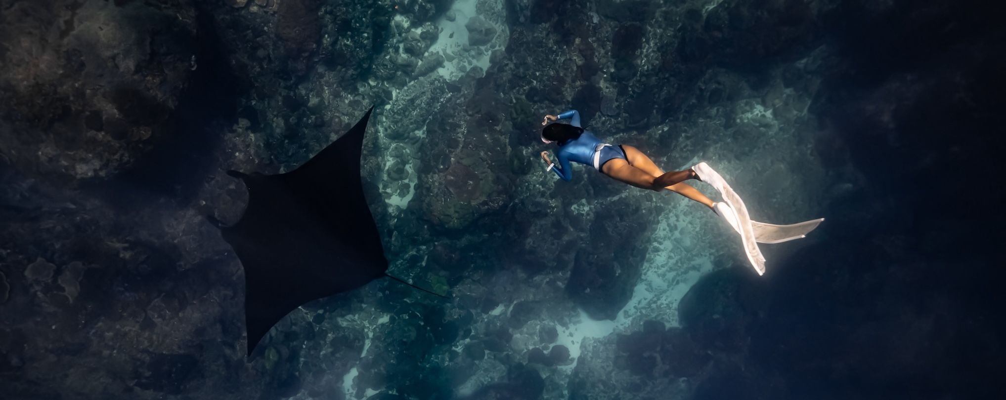 A freediver in white fins gliding alongside a manta ray over a dark reef