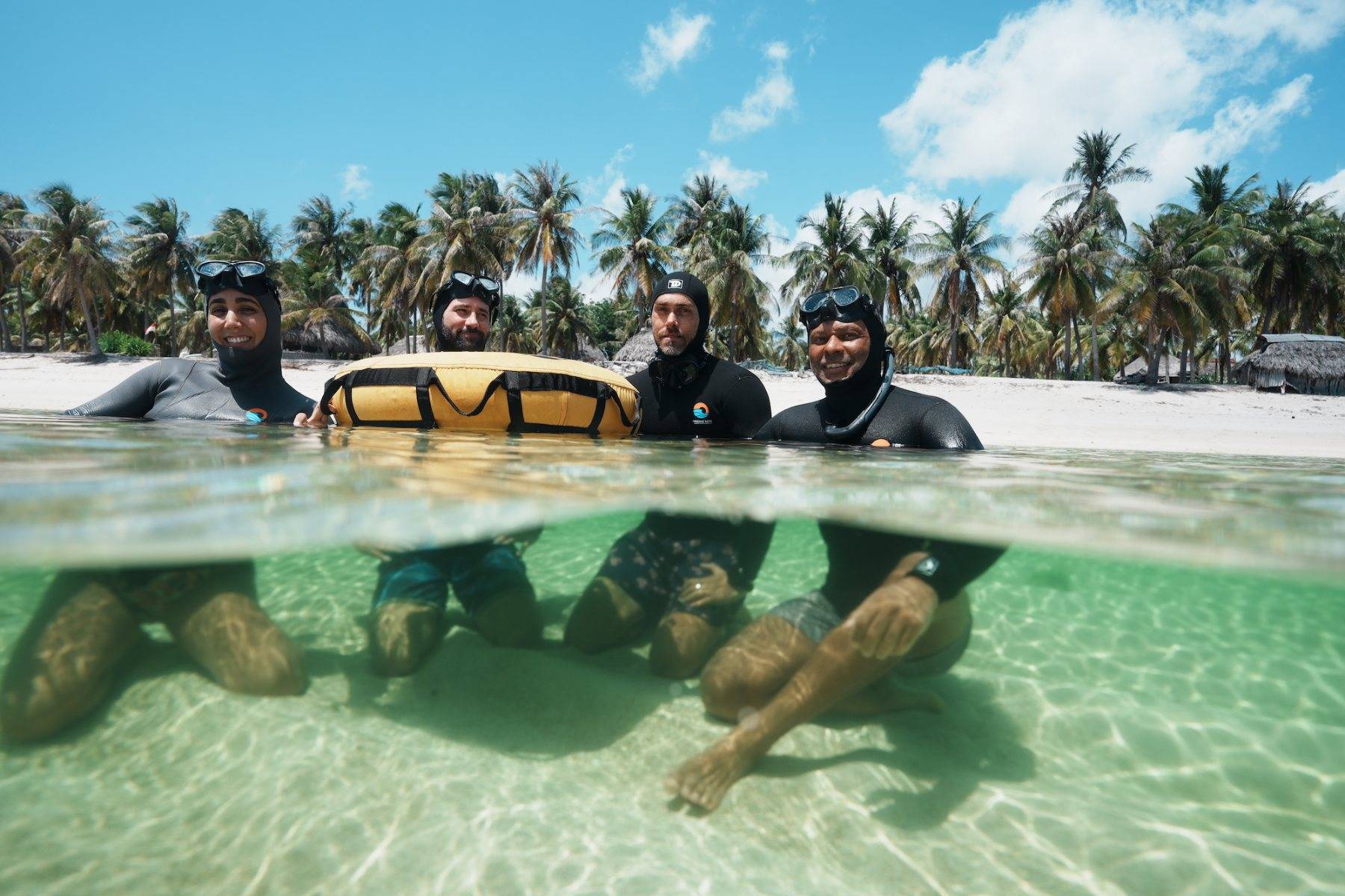 Four freedivers in hoods and masks smiling at the buoy, half above and half below the lagoon surface