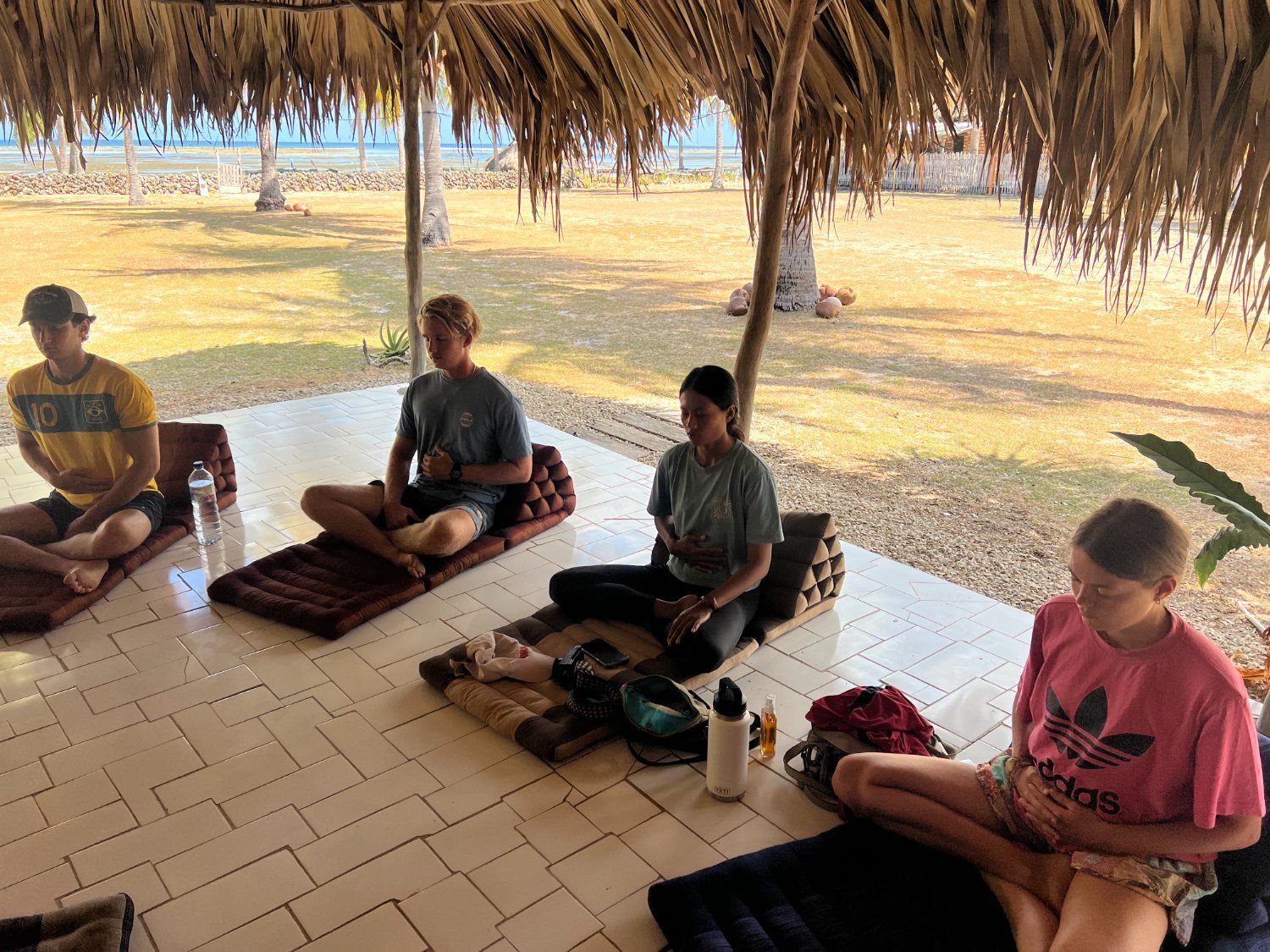 Students sitting in breathwork practice in the shala, the beach and lagoon glowing behind them