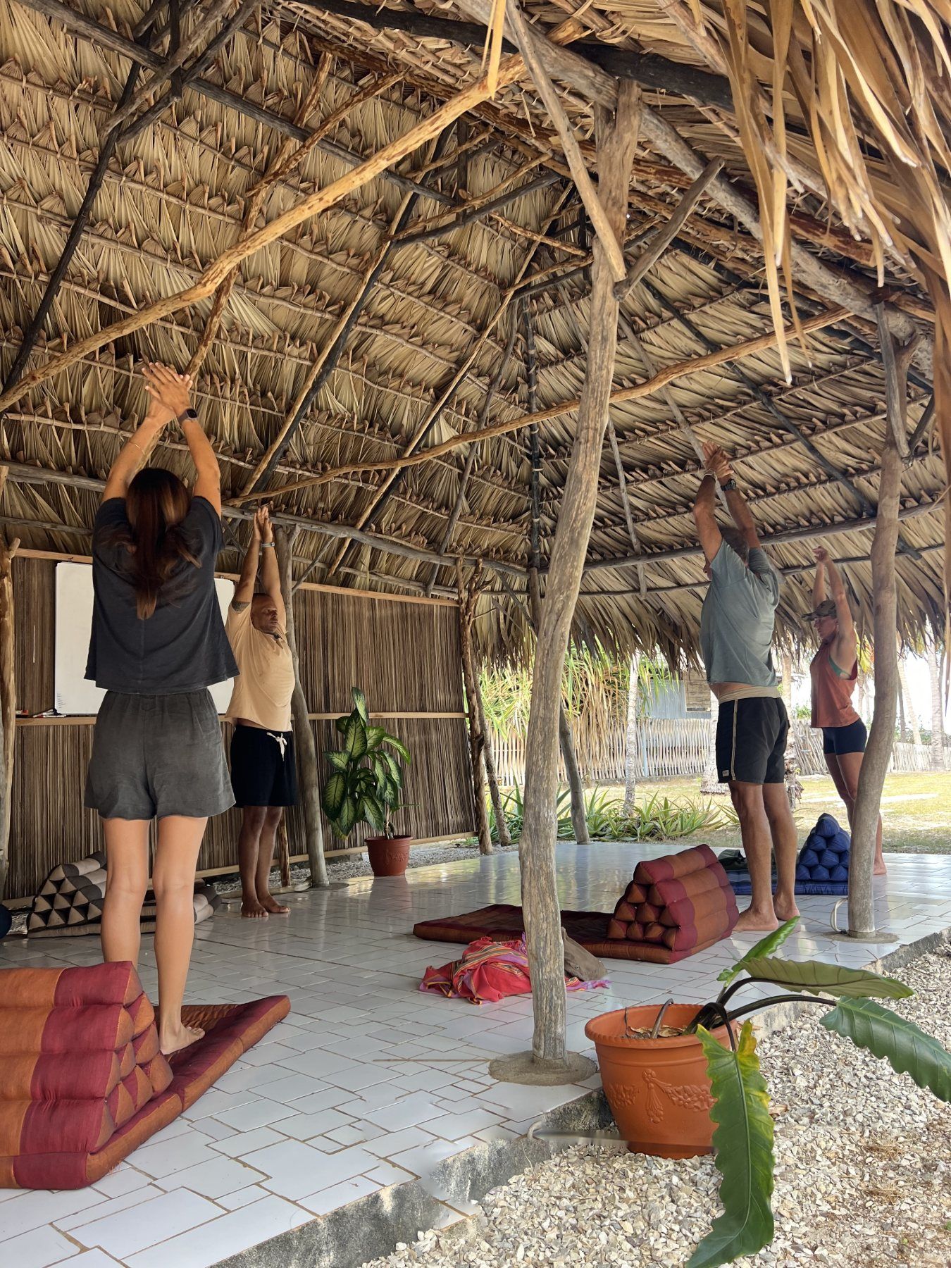 A group stretching arms overhead under the palm-thatch roof of the shala